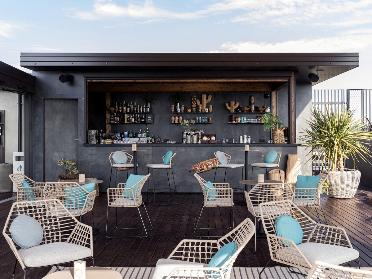 An open-fronted rooftop bar with a dark counter, stocked shelves and carved tiki figures, with woven armchairs and turquoise cushions on the timber deck in front of it.