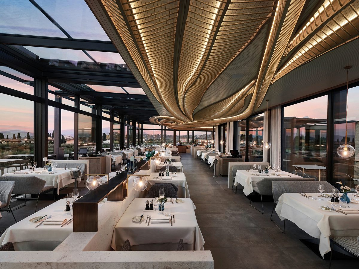 A long fine-dining room with white-clothed tables and grey armchairs beneath a sculptural gold ceiling, glazed along both sides and overhead, with sunset light over the countryside outside.
