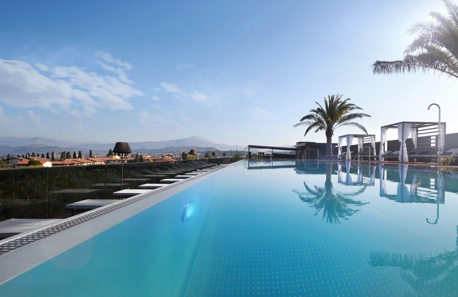The rooftop pool at Quellenhof Luxury Resort Lazise, its far edge level with the water, sun loungers along one side and curtained cabana beds and palms at the far end, above olive groves, tiled village roofs and distant hills.