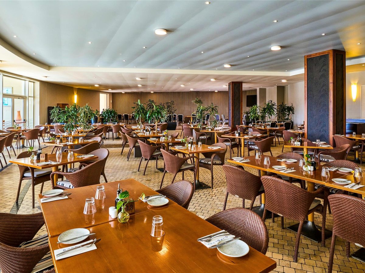 Large indoor restaurant with rows of wooden tables, woven brown chairs, potted palms and a curved white ceiling.