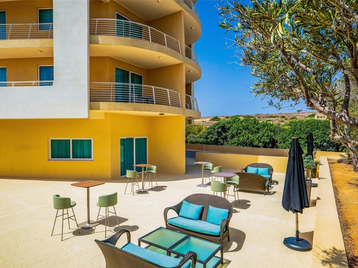 Paved terrace with wicker sofas and turquoise cushions, high tables and green stools at the foot of a yellow tower with curved balconies, an olive tree to one side.
