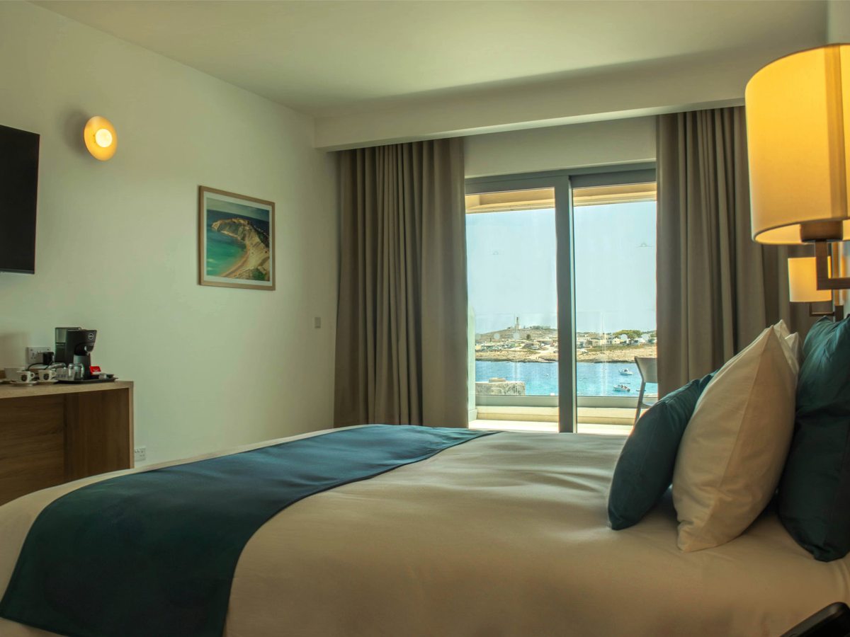 A double bed with teal and cream cushions, a coffee machine and cups on a side unit, and open curtains at sliding doors showing a low headland and blue sea.
