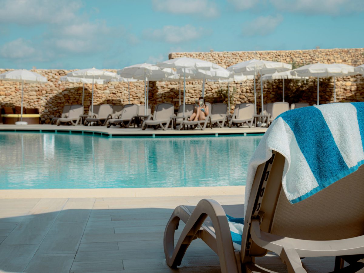 A large outdoor pool edged with sun loungers and white parasols in front of a long rubble-stone wall, with a blue and white striped towel draped over a lounger in the foreground.