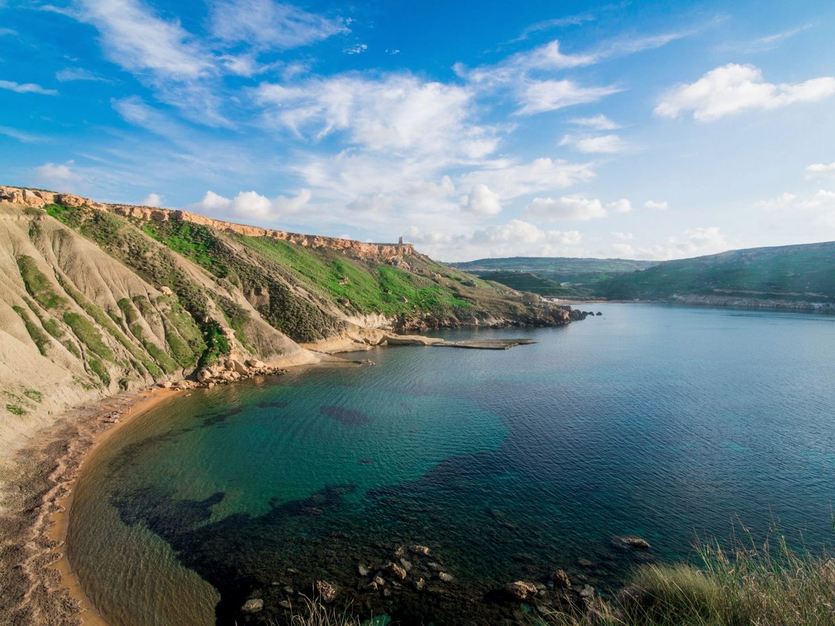 A curving bay with a strip of sand below steep pale clay slopes and green terraces, the shallows turning turquoise under a blue sky with scattered cloud.