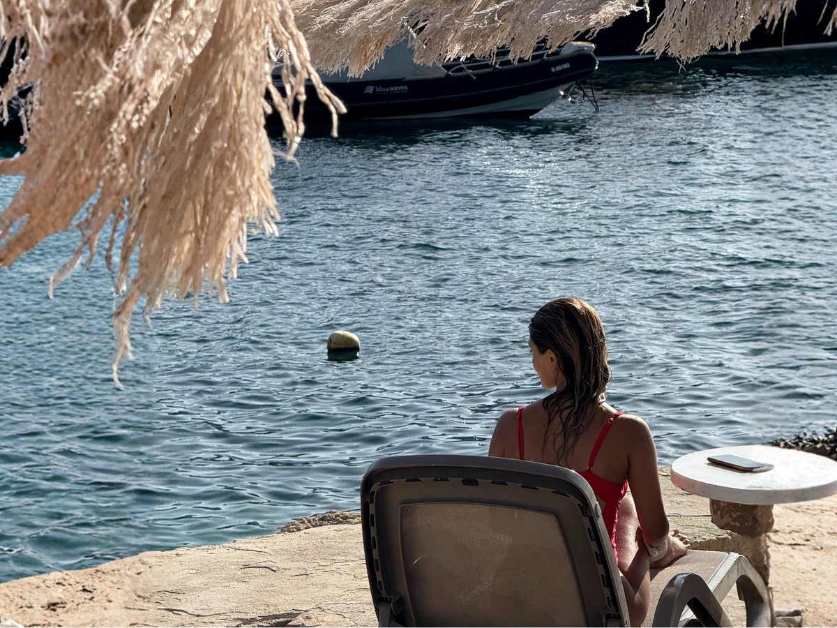 A sun lounger and a small round table on a stone terrace at the water's edge under a thatched shade, with a rigid inflatable boat moored against the rocks across the inlet.