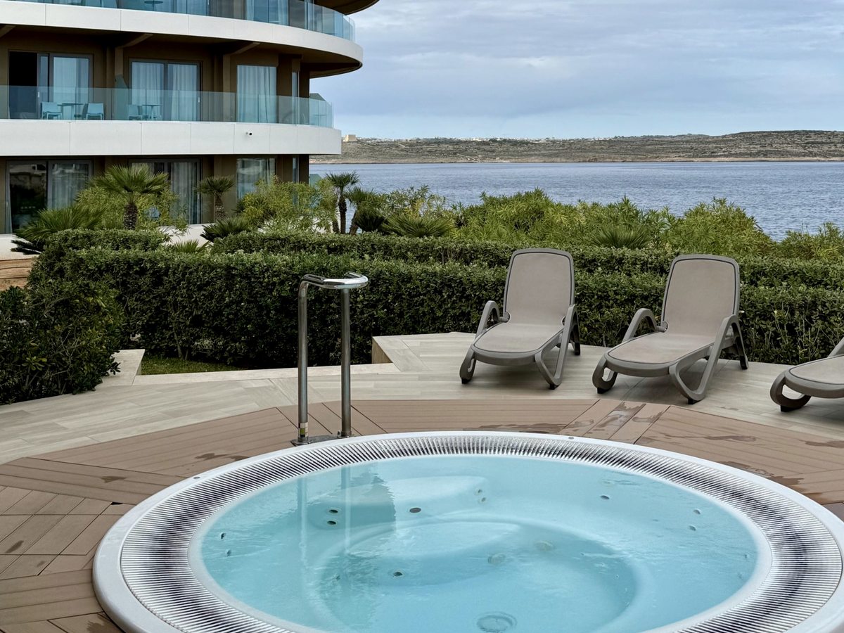 A circular jacuzzi set into a pale wooden deck with two sun loungers behind it, clipped hedges and palms, and the sea and a low coastline beyond.
