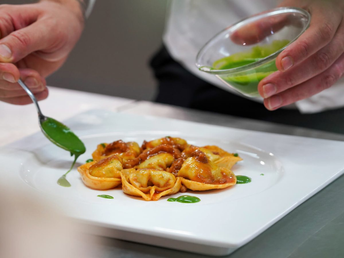 A chef's hands spooning green sauce over a plate of stuffed pasta on a square white plate.