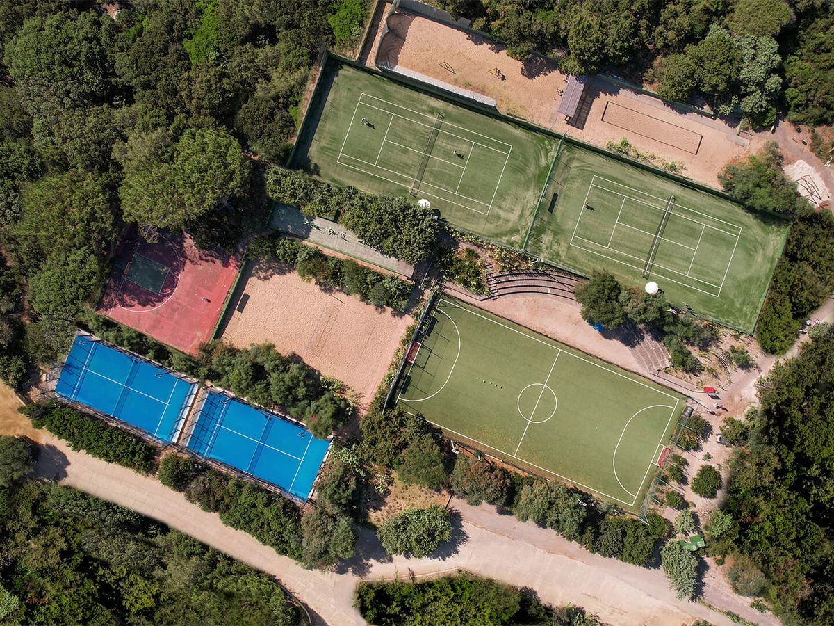 Aerial view of enclosed blue courts, a red court and a green tennis court beside a green five-a-side football pitch, ringed by pine and juniper woodland.