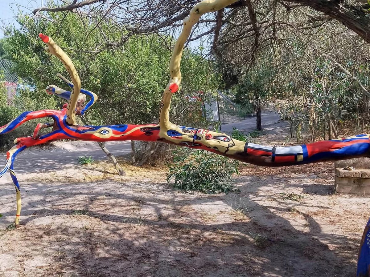 A fallen tree trunk painted in red, blue and cream patterns standing as a sculpture beside a sandy path among olive trees and scrub.