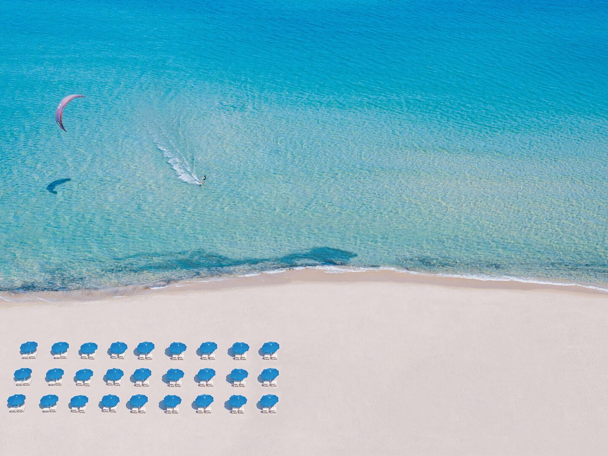 Aerial view of three neat rows of blue parasols and loungers on pale sand, with clear turquoise water and a kitesurfer offshore.