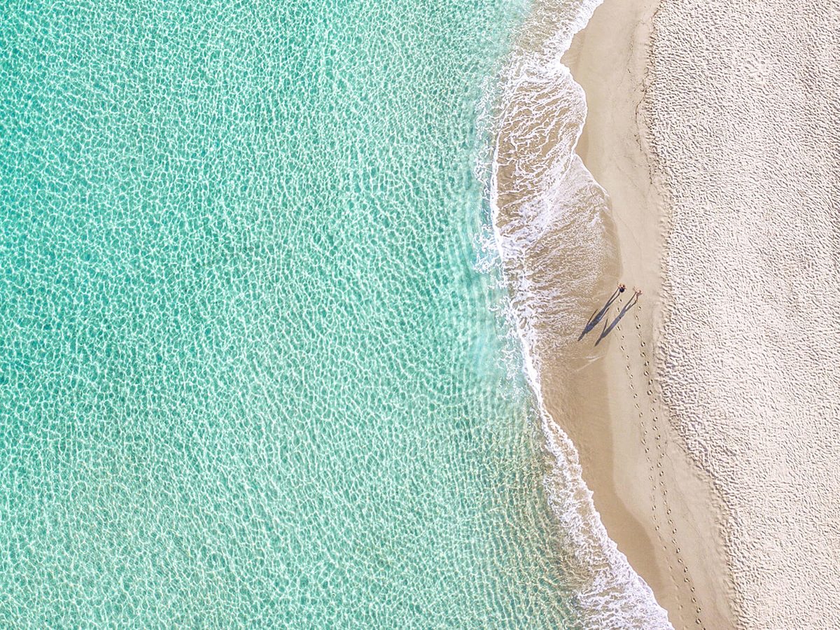 Aerial view of clear turquoise shallows breaking onto a wide strip of pale sand, with two people walking at the water's edge.