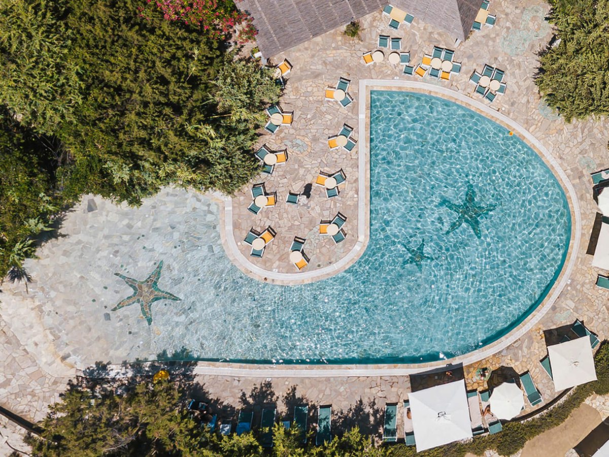 Aerial view of a horseshoe-shaped pool wrapped around a stone sun terrace, with starfish mosaics in the paving and green loungers and parasols around it.