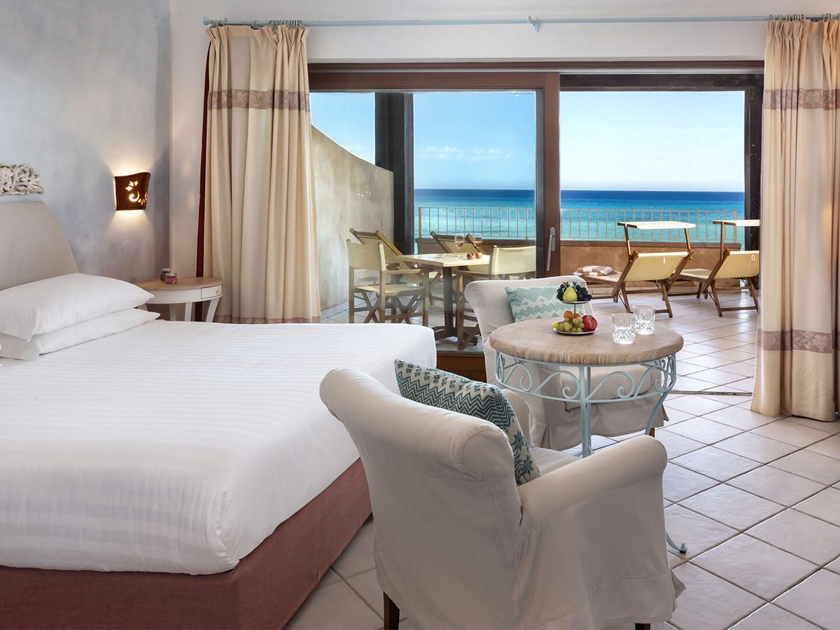 Guest room with a double bed, two pale armchairs and a small round table on a tiled floor, with open doors onto a furnished terrace and the sea behind.