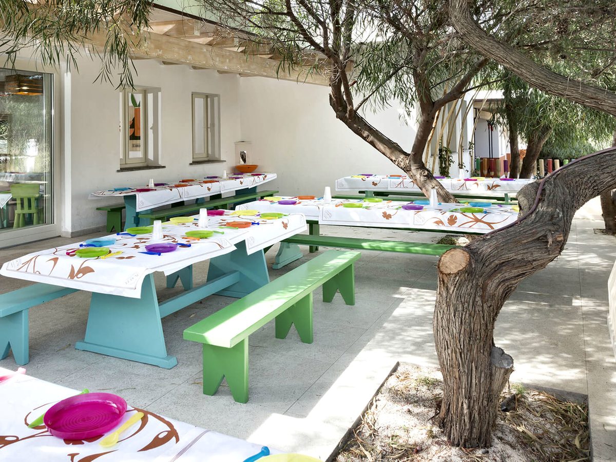 Child-height picnic tables with green and blue benches and bright plastic plates on a shaded terrace beside a white building, under an olive tree.