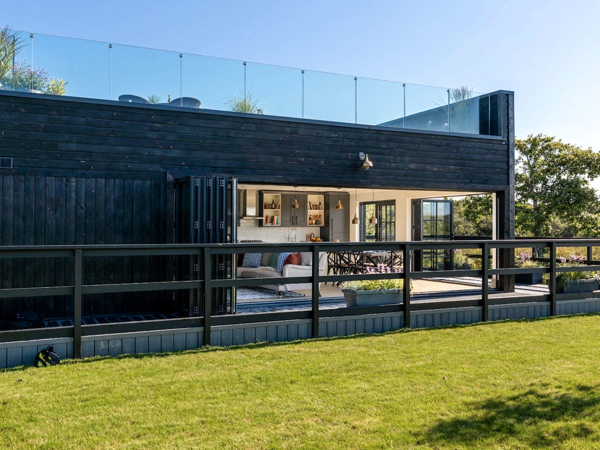 A single-storey black timber lodge with folding glass doors open onto a decked terrace and a glass-balustraded roof terrace planted with grasses above.