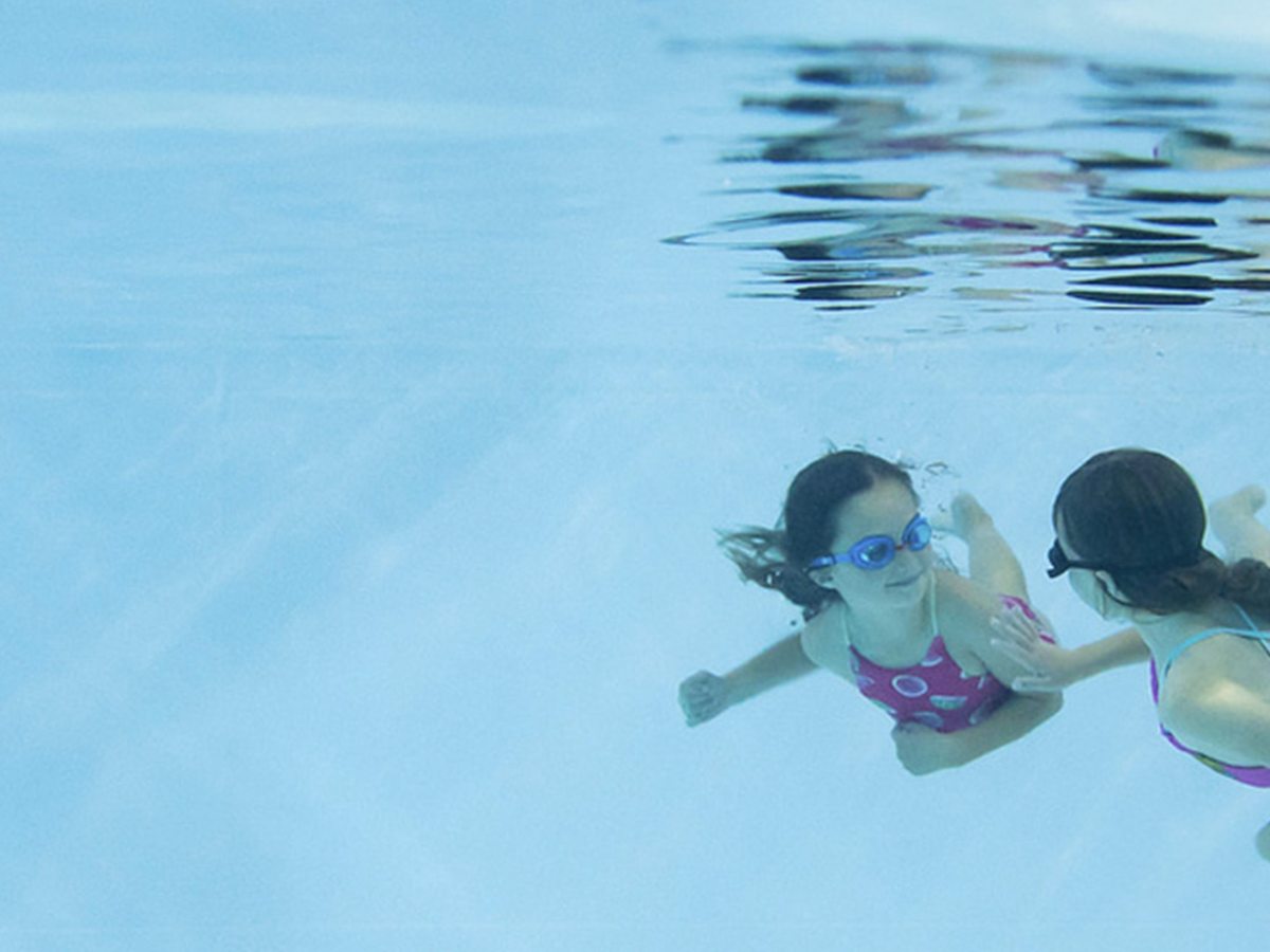 Two girls in goggles swimming underwater in a pale blue indoor pool.