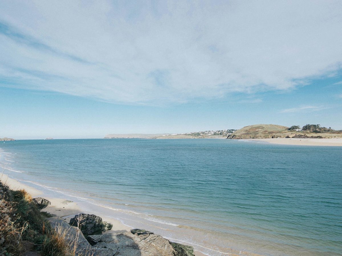 A wide sandy bay with pale turquoise water, rocks in the foreground and a grassy headland and scattered houses across the water.