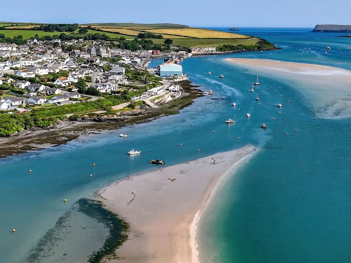 An aerial view of a wide blue estuary with pale sandbanks, moored boats and a village of white houses along the shore.