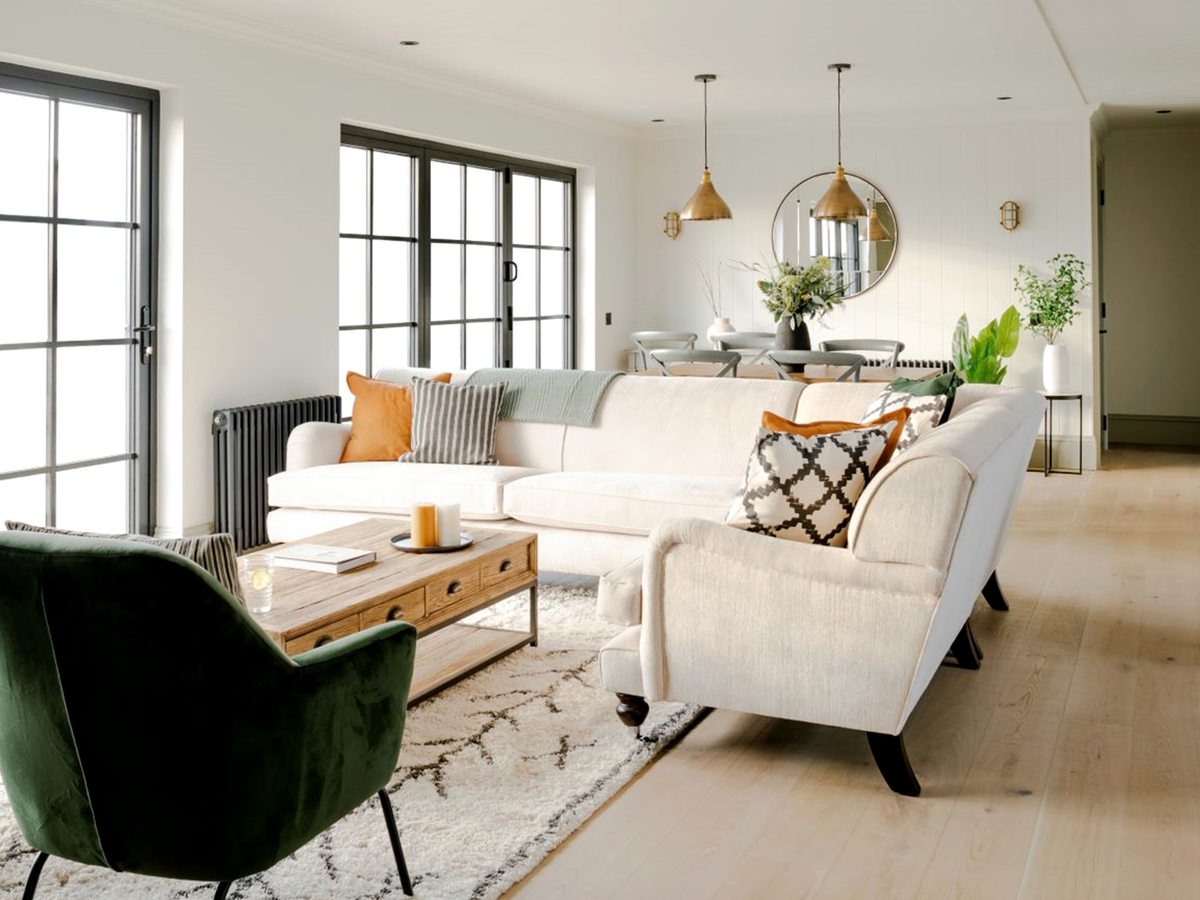 A bright lounge with two cream sofas, a green velvet armchair and a wooden coffee table on a pale rug, with a dining table and brass pendants at the far end.