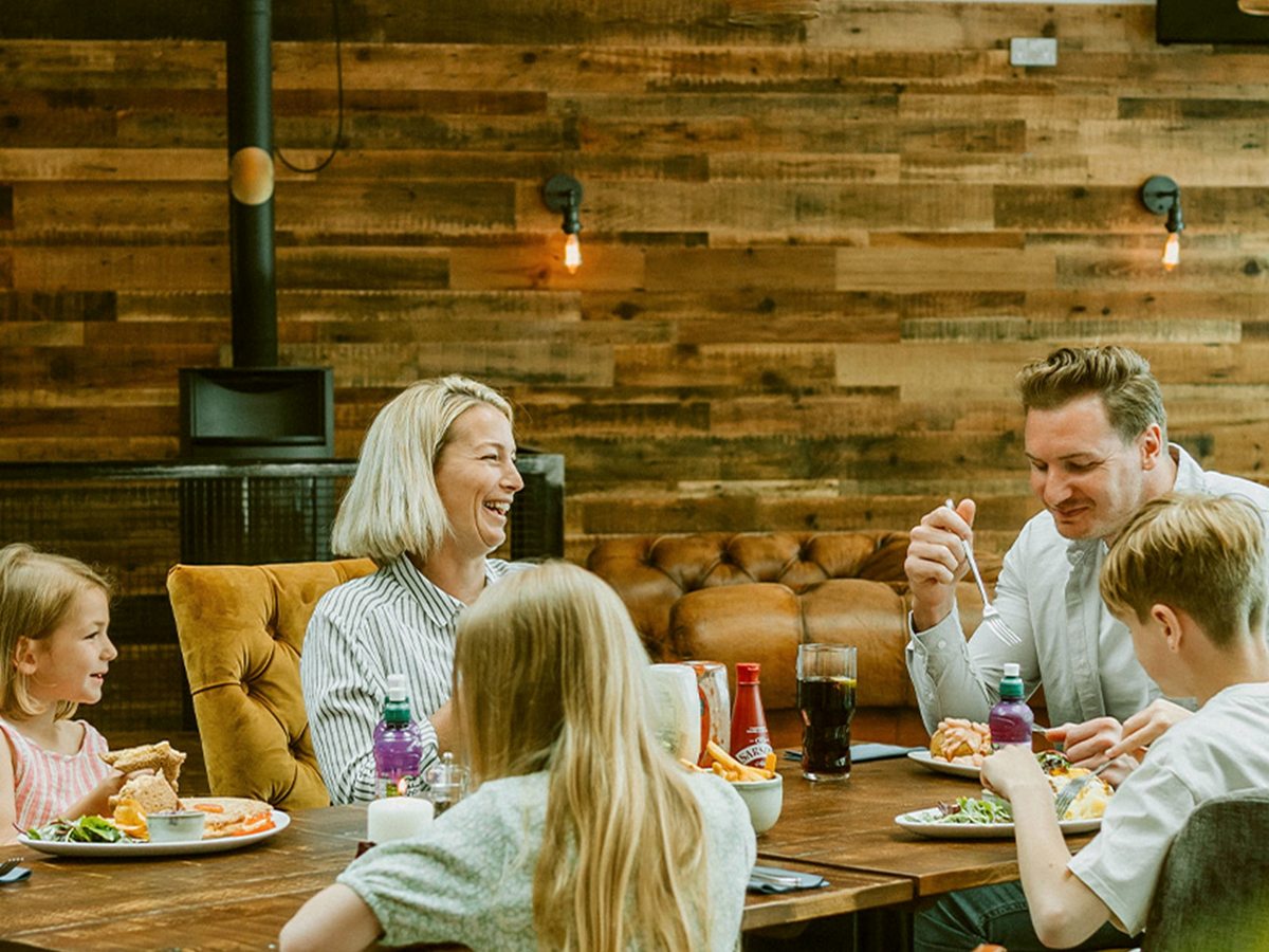Two adults and three children sitting round a long wooden table eating and laughing, against a reclaimed-timber wall with a wood-burning stove behind.