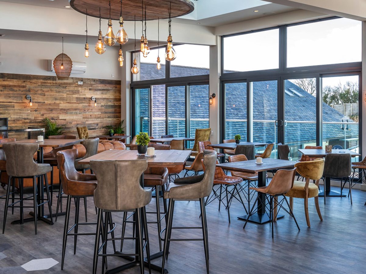 A restaurant interior with reclaimed-timber walls, leather high stools at tall tables, low tan and mustard chairs, and a cluster of filament pendant lights under a full-height glazed wall.