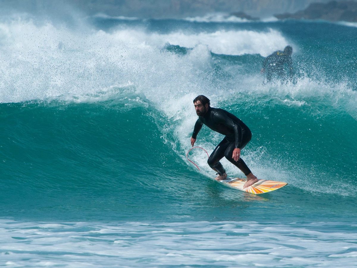 A surfer in a black wetsuit riding along the face of a breaking turquoise wave with white spray behind.