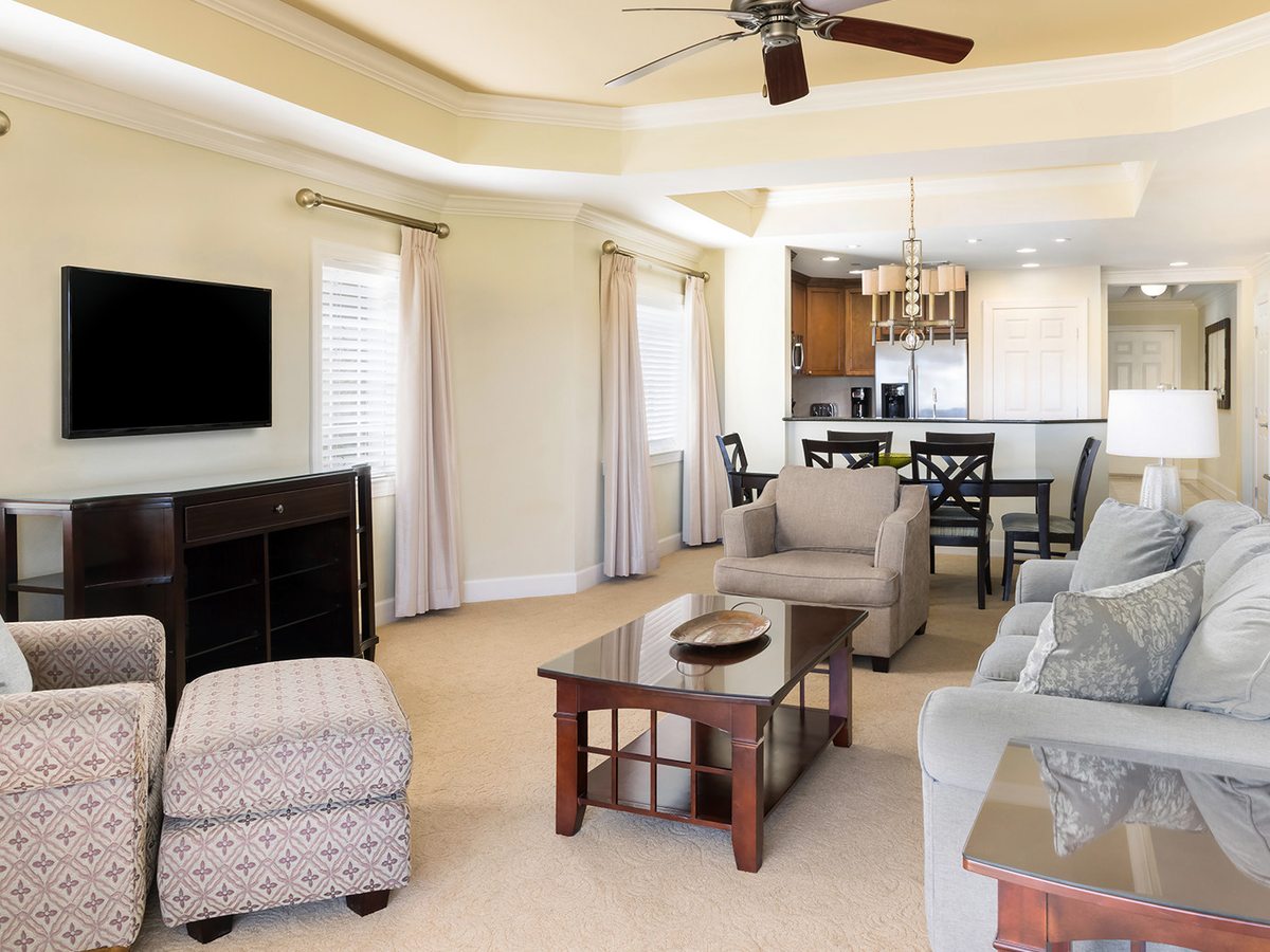 An open-plan villa interior with two sofas and an armchair facing a wall-mounted television, a dining table behind and an open kitchen at the back.