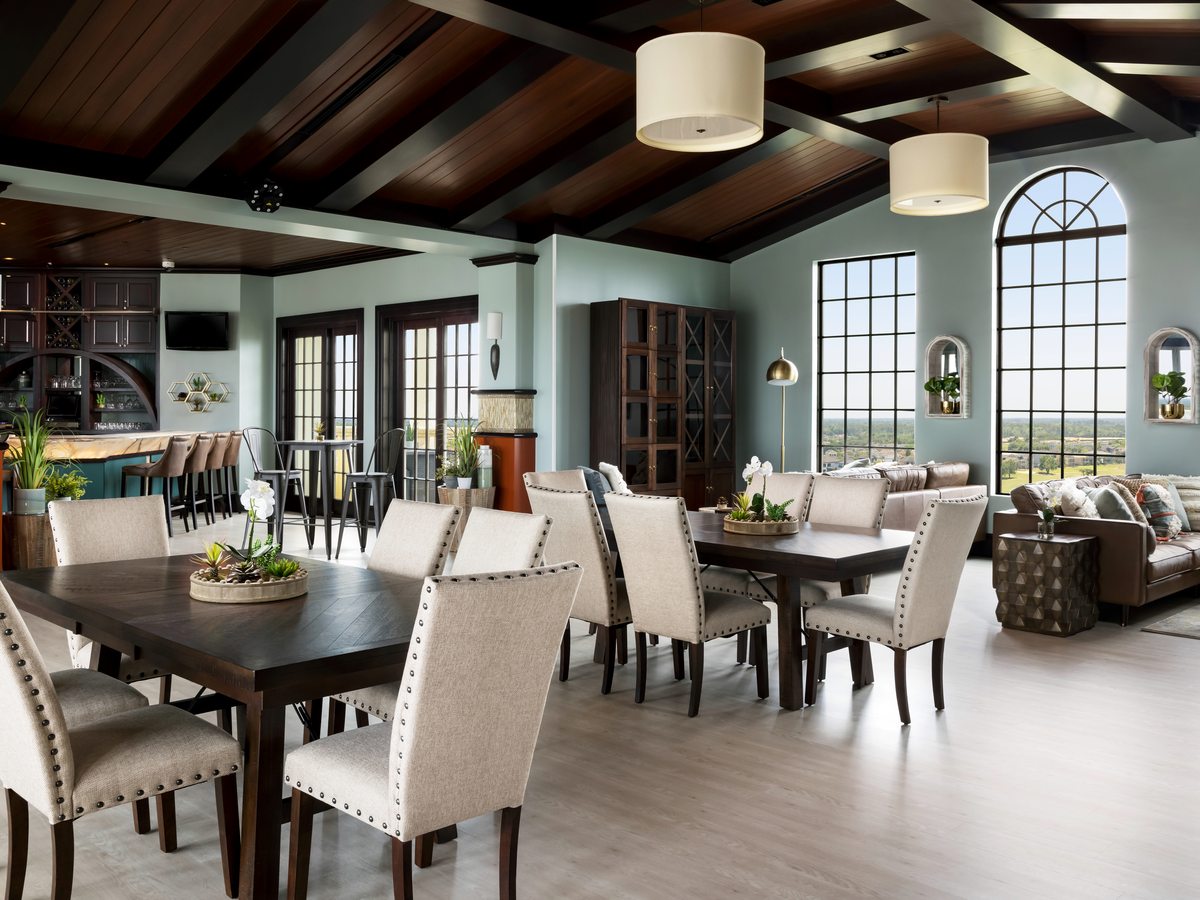 A villa dining area with a long table and many upholstered chairs under a beamed timber ceiling, a breakfast bar with stools and a lounge beyond.