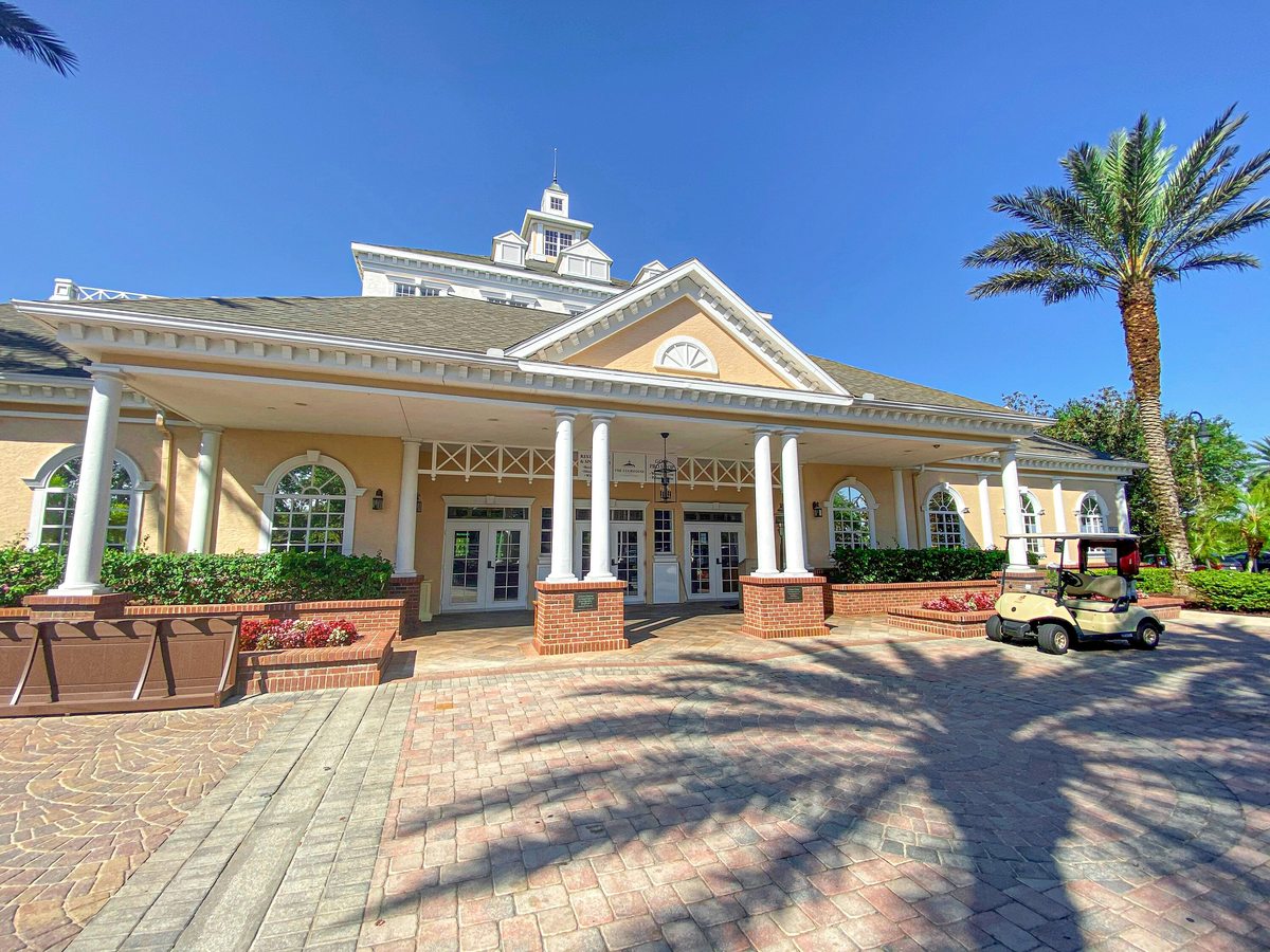 A columned clubhouse building with a classical pediment and cupola, a brick-paved forecourt and a golf cart parked at the entrance among palm trees.