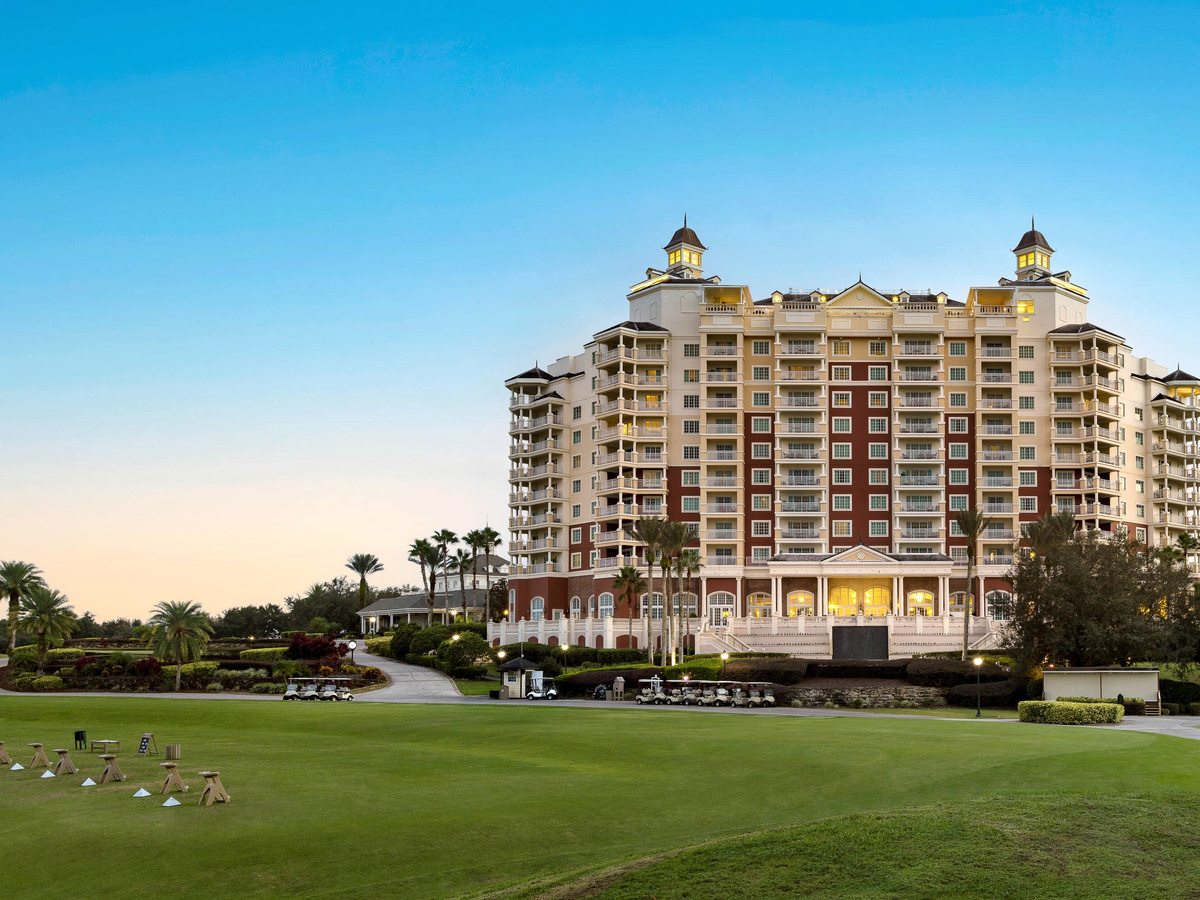 A tall cream resort tower with twin rooftop cupolas rising above manicured lawns and palm trees at dusk, its windows lit.