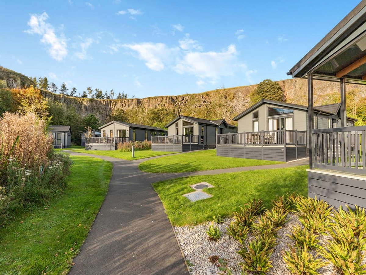 Rows of grey timber lodges with railed verandas beside a curving tarmac path through mown lawns, with high limestone quarry cliffs and blue sky behind.