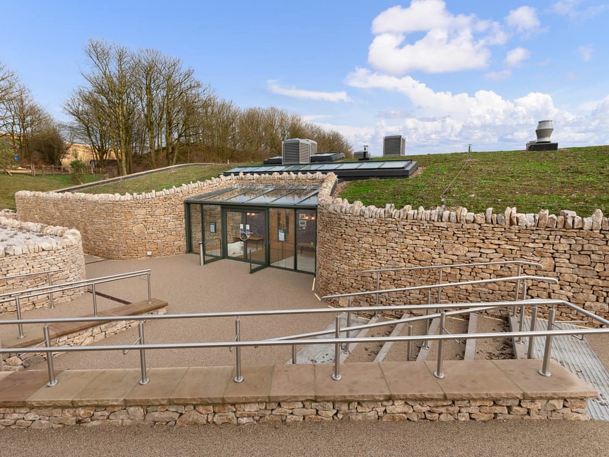 A glazed entrance set into a grass-roofed building between dry-stone walls, reached by a wide paved ramp with metal handrails running alongside a flight of steps.
