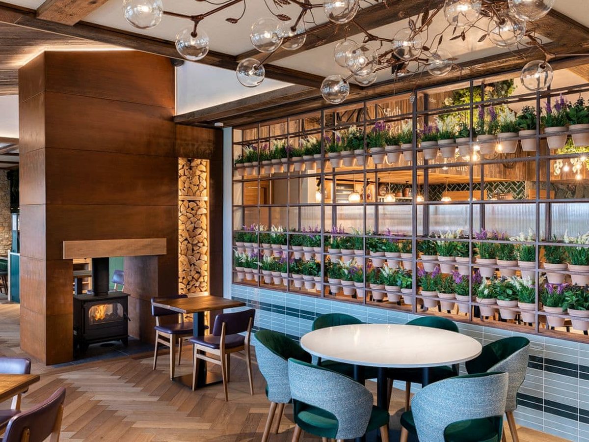 Restaurant tables beside a full-height metal frame filled with rows of potted plants, next to a wood-burning stove in a copper-clad chimney with stacked logs.