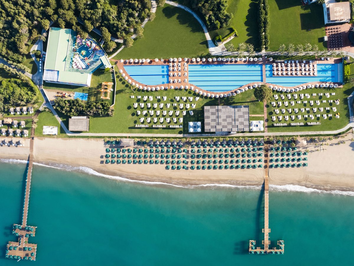 An aerial straight down over the beachfront: rows of green parasols on the sand, two piers over turquoise water, and the long pools, sunbeds and lawns behind.