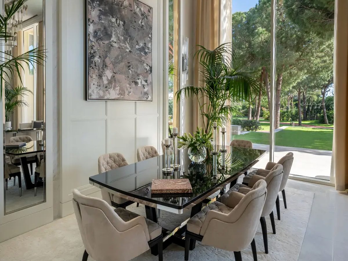 A villa dining room with a long black glass table and eight upholstered chairs in front of floor-to-ceiling windows onto a lawn.