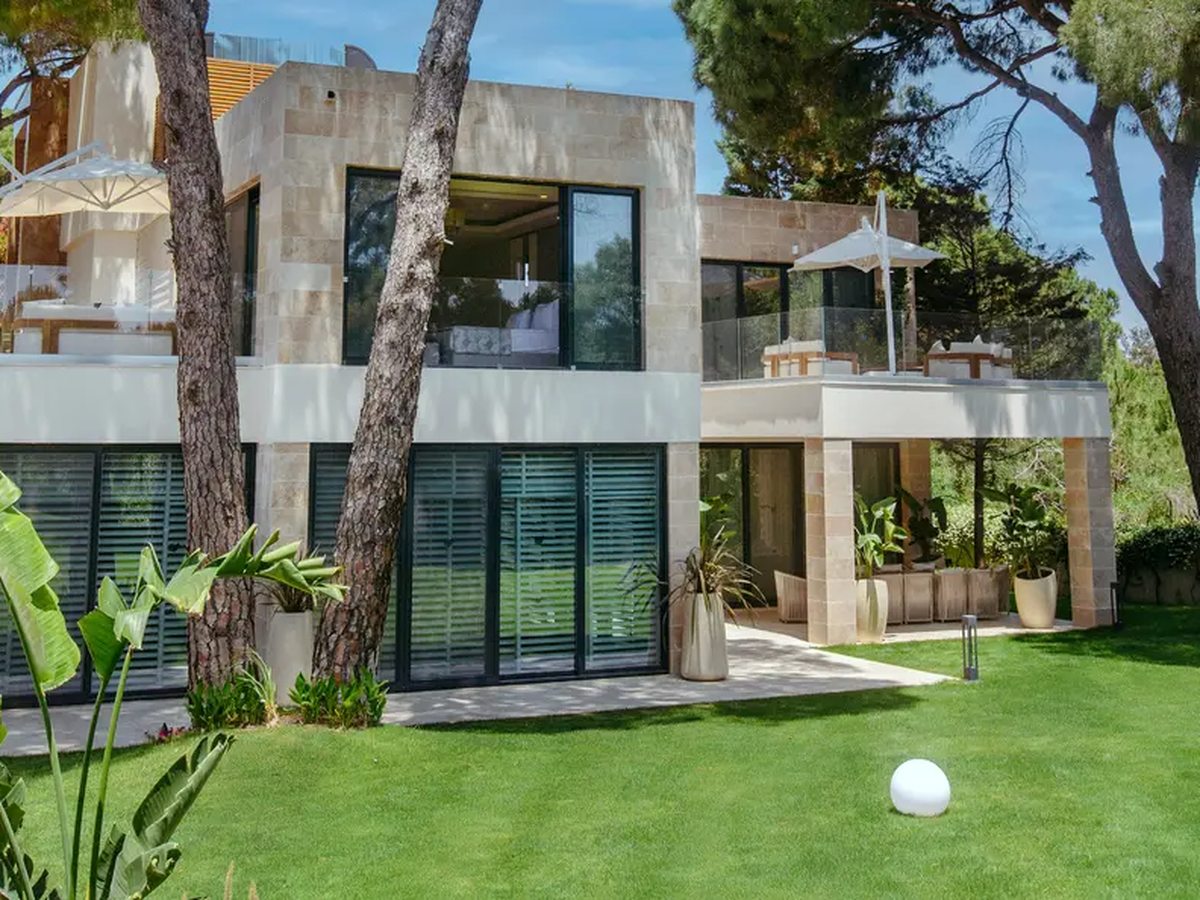 A two-storey stone-and-glass villa with a first-floor terrace and full-height sliding doors, standing on a lawn among umbrella pines.