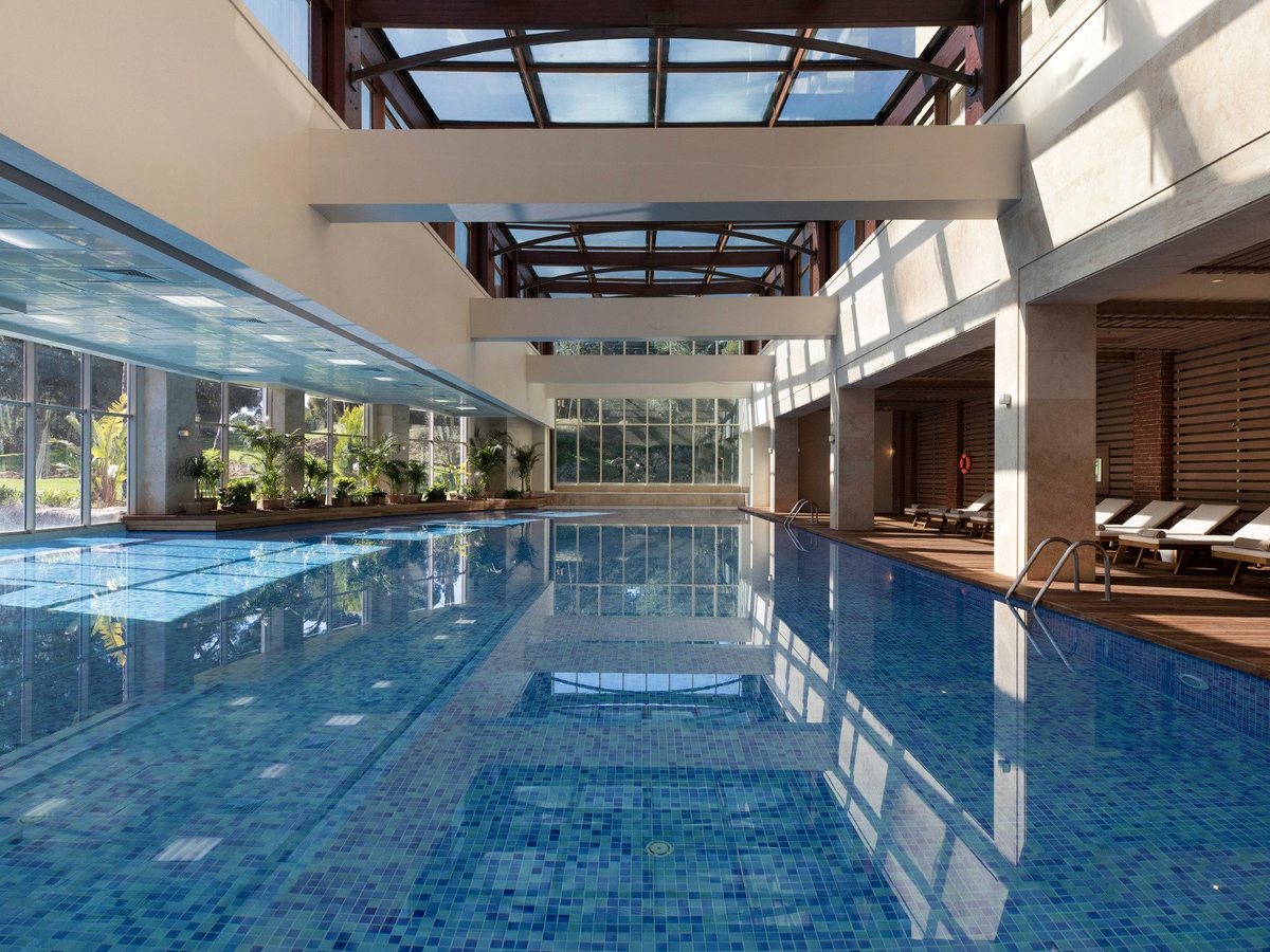 A long indoor pool tiled in blue mosaic under a glazed roof, with glass walls onto the gardens and loungers along one side.