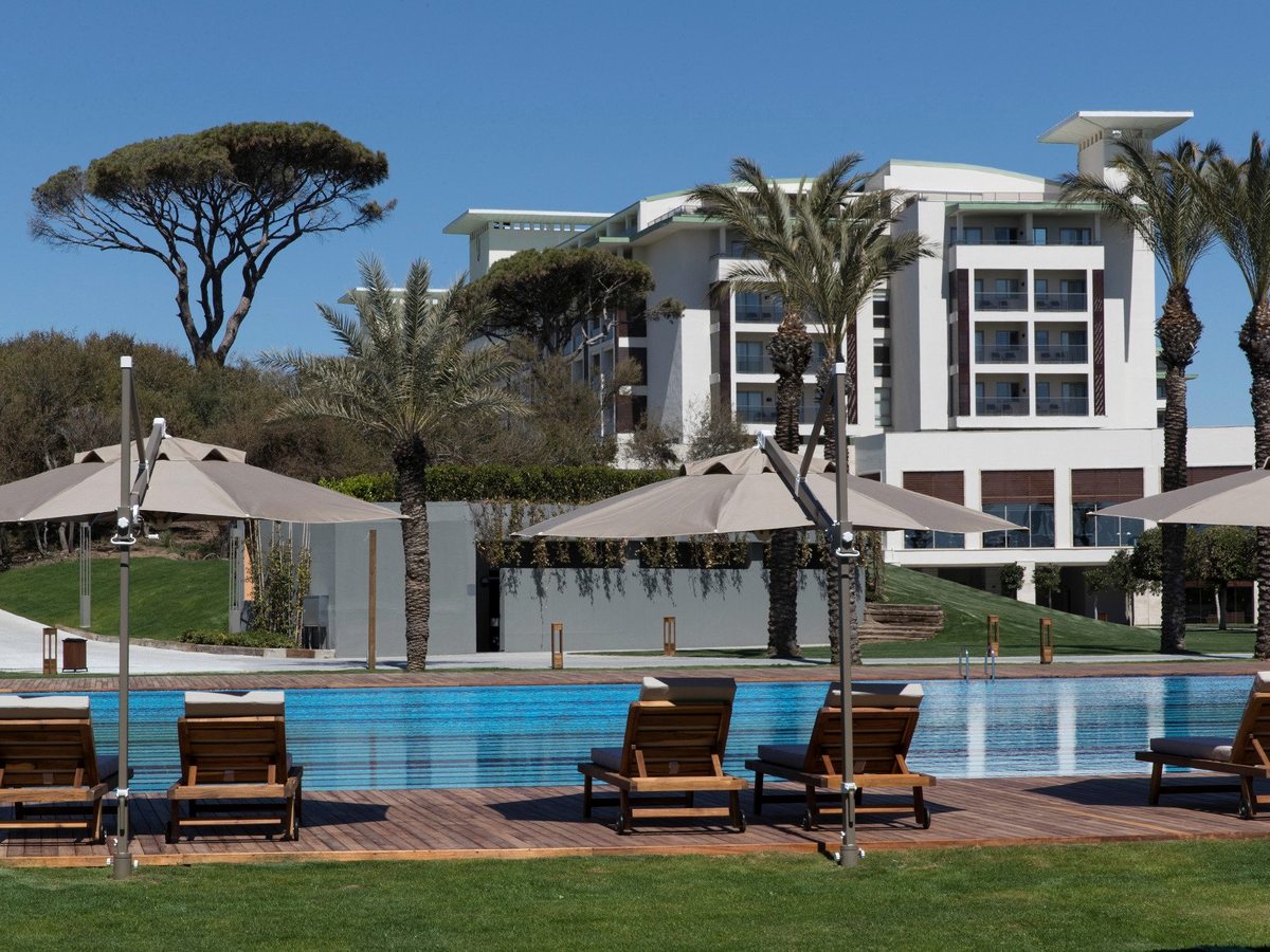 A long pool edged with a timber deck, loungers and parasols, with lawns, palms and the tall main hotel wing rising behind.