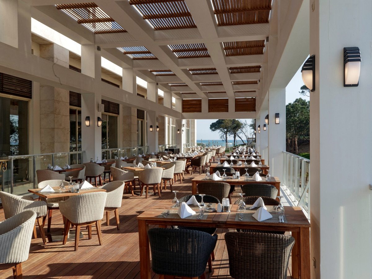 An open-sided restaurant terrace under a slatted timber pergola, with laid wooden tables, woven chairs and the sea visible at the far end.