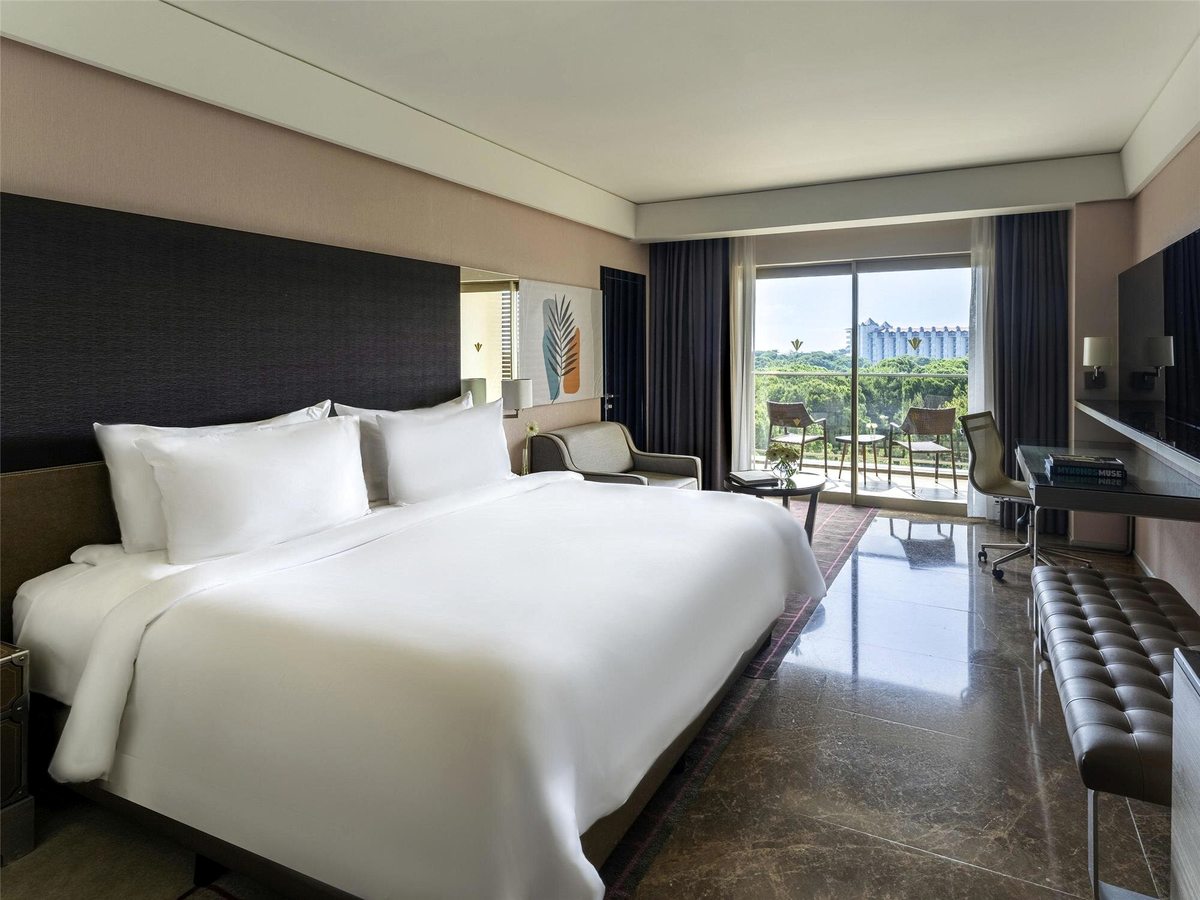 A king room with a dark padded headboard, white bedding, a bench at the foot of the bed, a desk along one wall and a balcony with chairs above pine woodland.