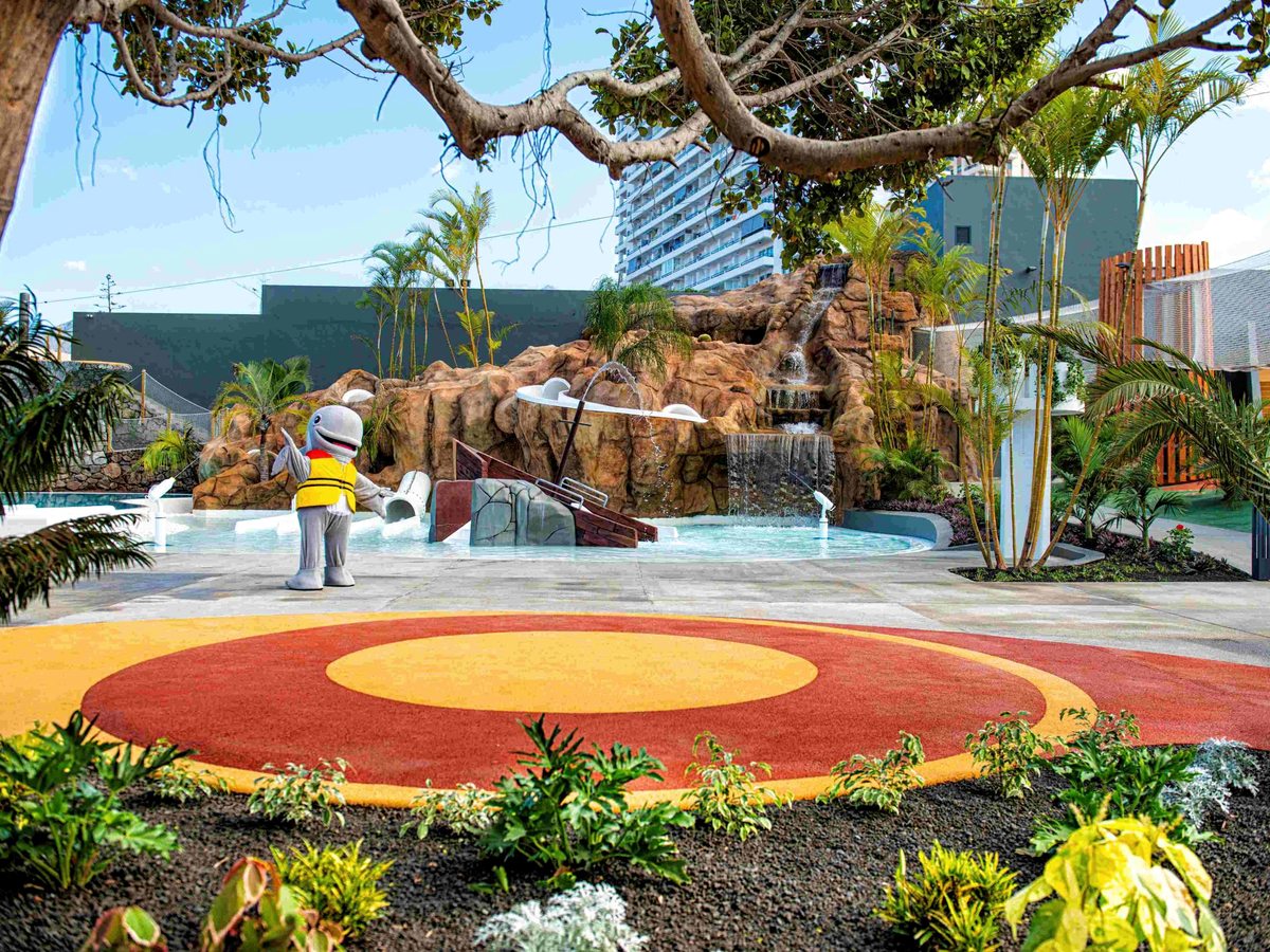 A children's water area with a sandstone rock waterfall, shallow pools, a wrecked wooden ship feature and a costumed grey whale mascot, with a red and yellow circular play surface in front.