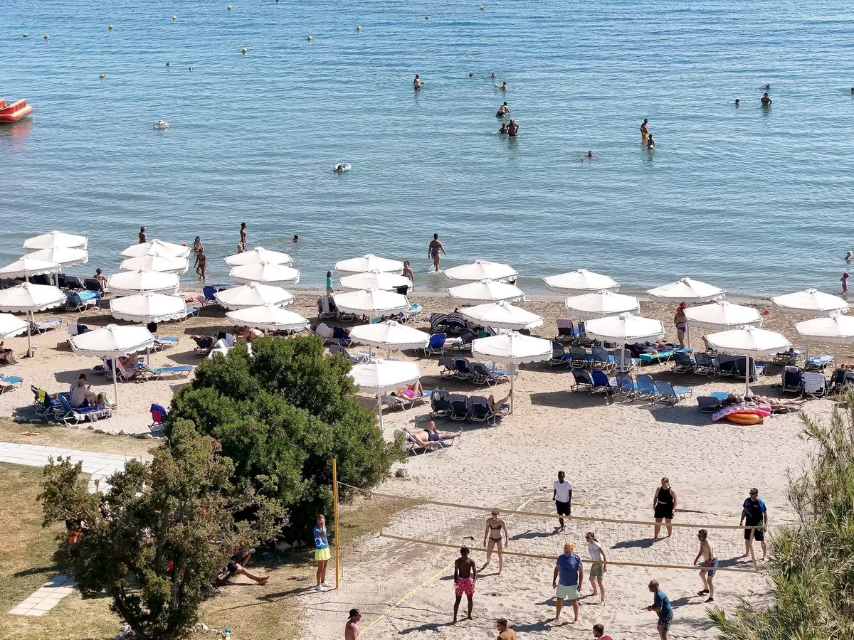 Rows of white parasols and sunbeds on the sand with people swimming in calm shallow water beyond, and a sandy volleyball court in the foreground.