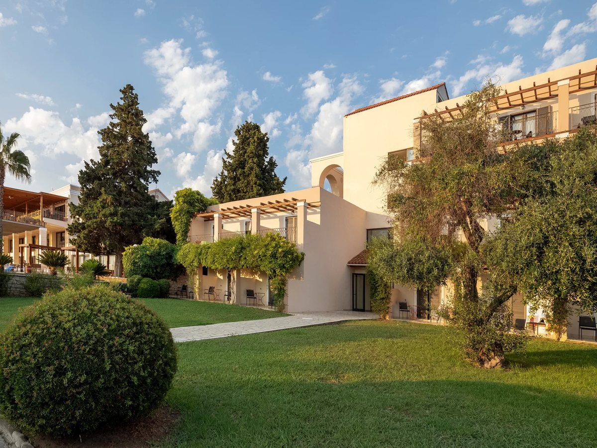 Lawns, clipped shrubs and mature trees between low cream buildings under a blue sky.