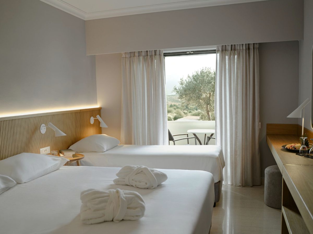 A room with a double bed and a single bed dressed in white with folded robes on top, a slatted wood headboard, and a window and balcony door looking onto olive trees.