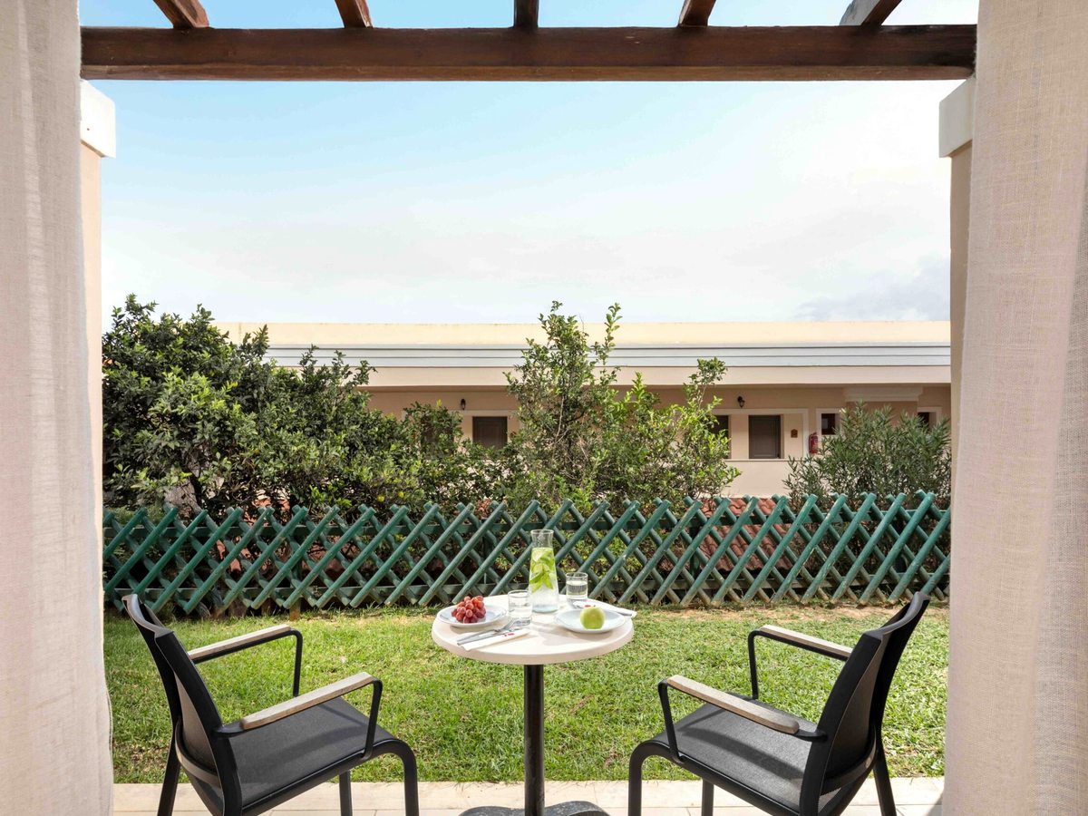 A ground-floor terrace under a timber pergola with two black chairs and a small table set with fruit and water, looking onto a lawn and a low cream building behind a trellis fence.