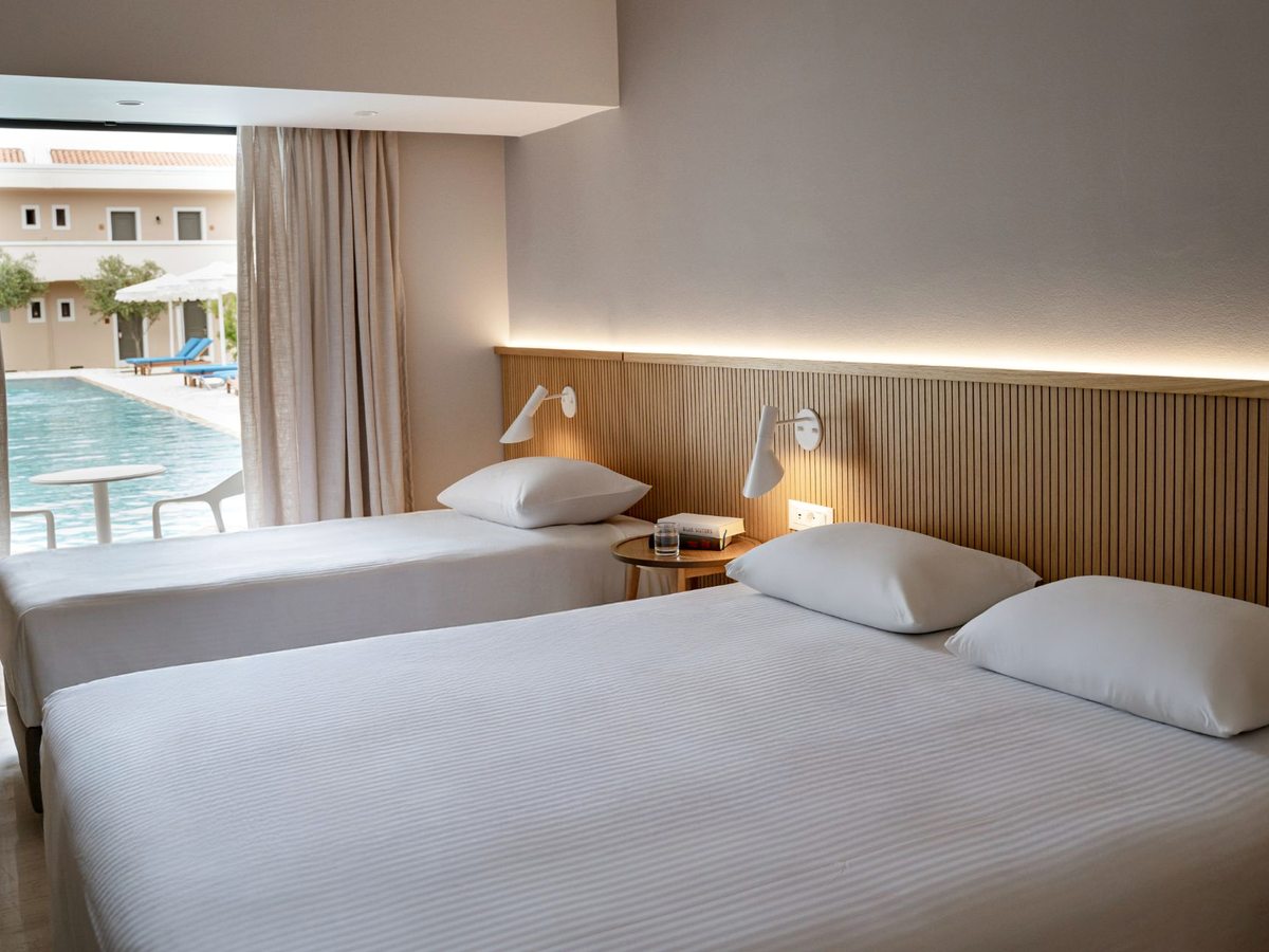 A room with two beds beside a wide opening onto a paved terrace at the very edge of a pool, with blue loungers and parasols on the far side.