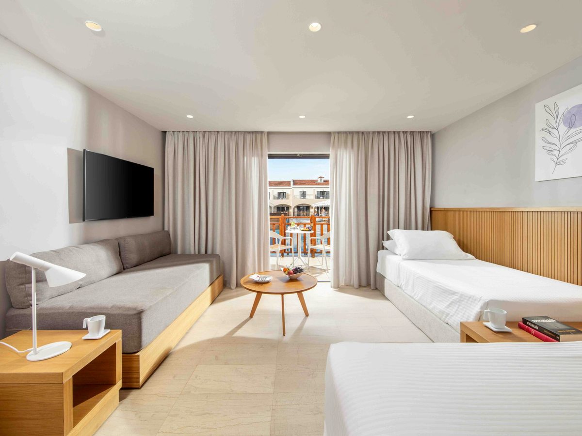 A large room with a grey sofa bed along one wall, two single beds opposite, a round wooden coffee table between them and glazed doors to a balcony set with a table and stools.