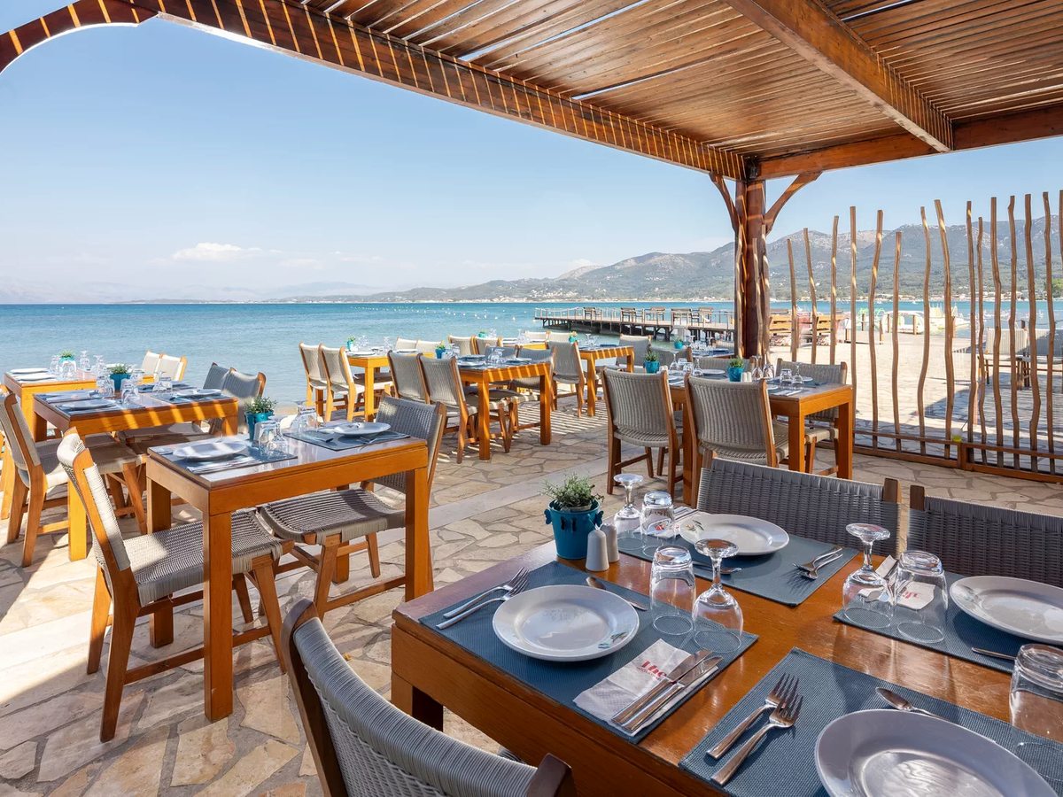 A restaurant under a wooden canopy on a stone floor beside the sand, its tables laid with blue mats, plates and glassware, with the resort's wooden pier and distant mountains behind.