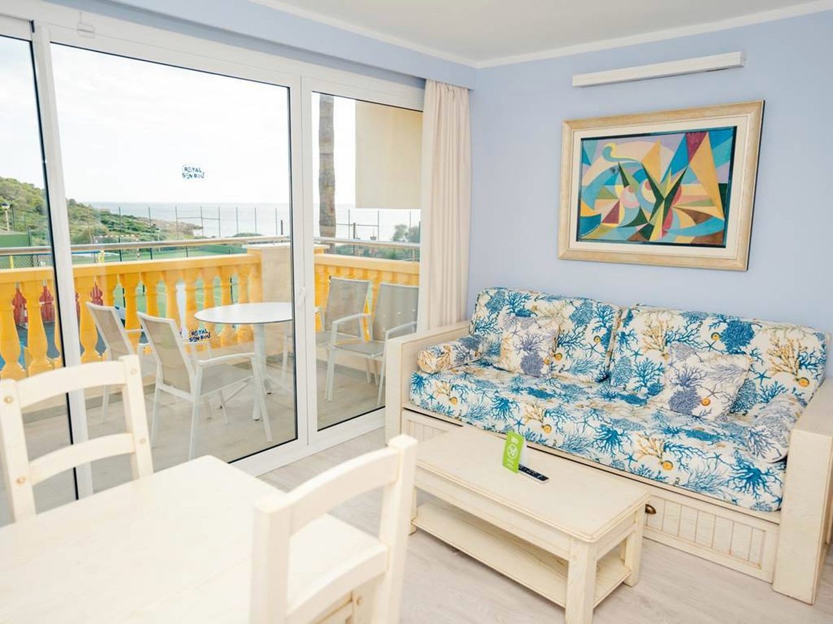An apartment living area with a blue coral-patterned sofa bed, a pale wooden coffee table and dining chairs, and sliding doors on to a balcony with yellow railings and the sea beyond.
