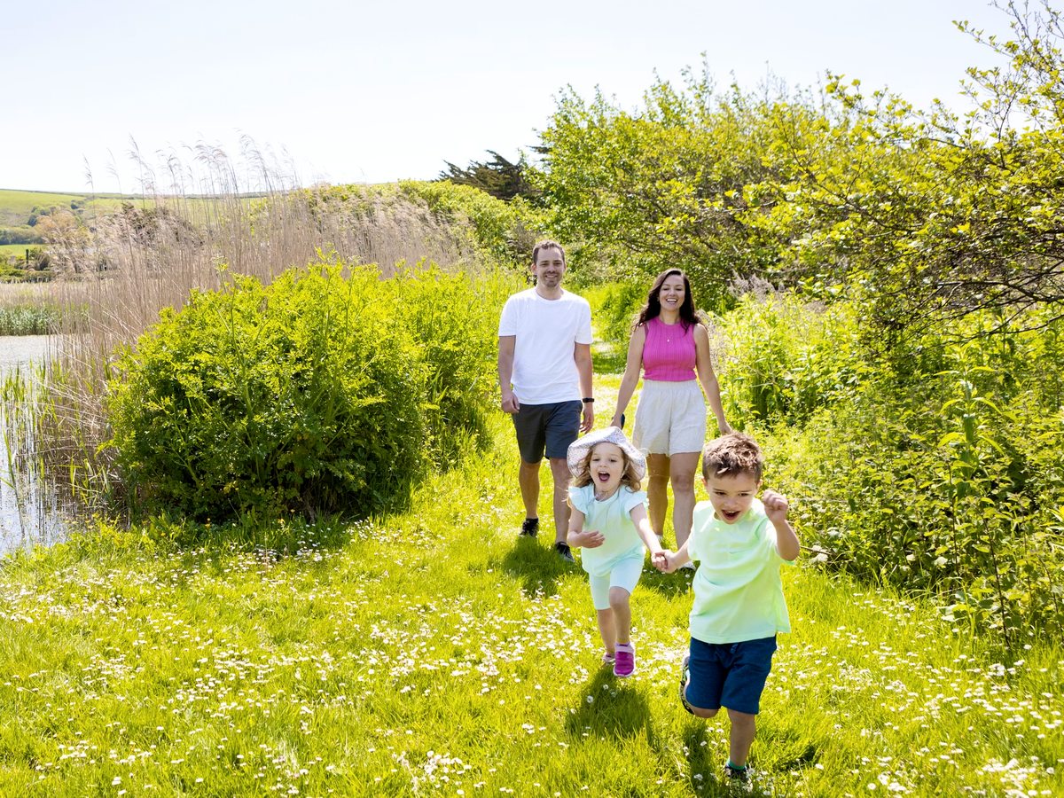 Two parents and two young children walking a grass path between hedgerow and reed beds beside a lake, with green hills in the distance.